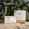 A box of Orique Liposomal Vitamin C with Zinc and Citrus Bioflavonoids is displayed outdoors with floral elements in the background.